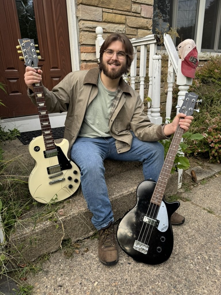 Chase seated on porch steps holding an electric guitar and a bass guitar