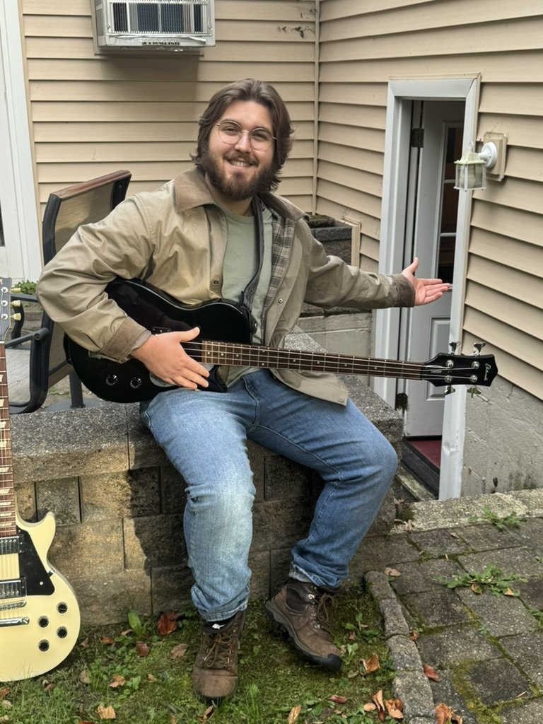Chase smiling and gesturing toward the studio entrance with a bass guitar on his lap