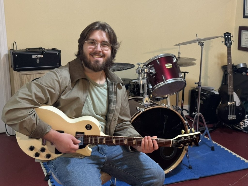 Chase smiling with an electric guitar in his home studio, with a drum kit and amps behind him