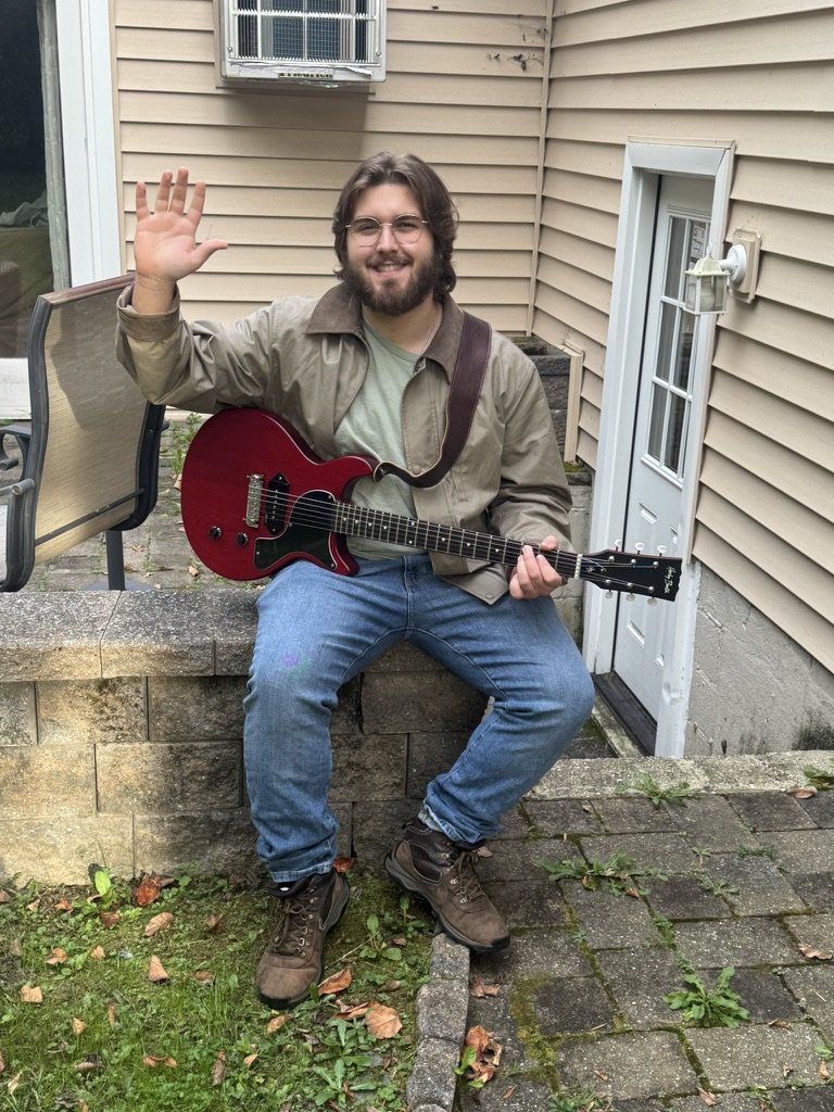 Chase waving hello while holding a red electric guitar on a stone wall outside the studio door